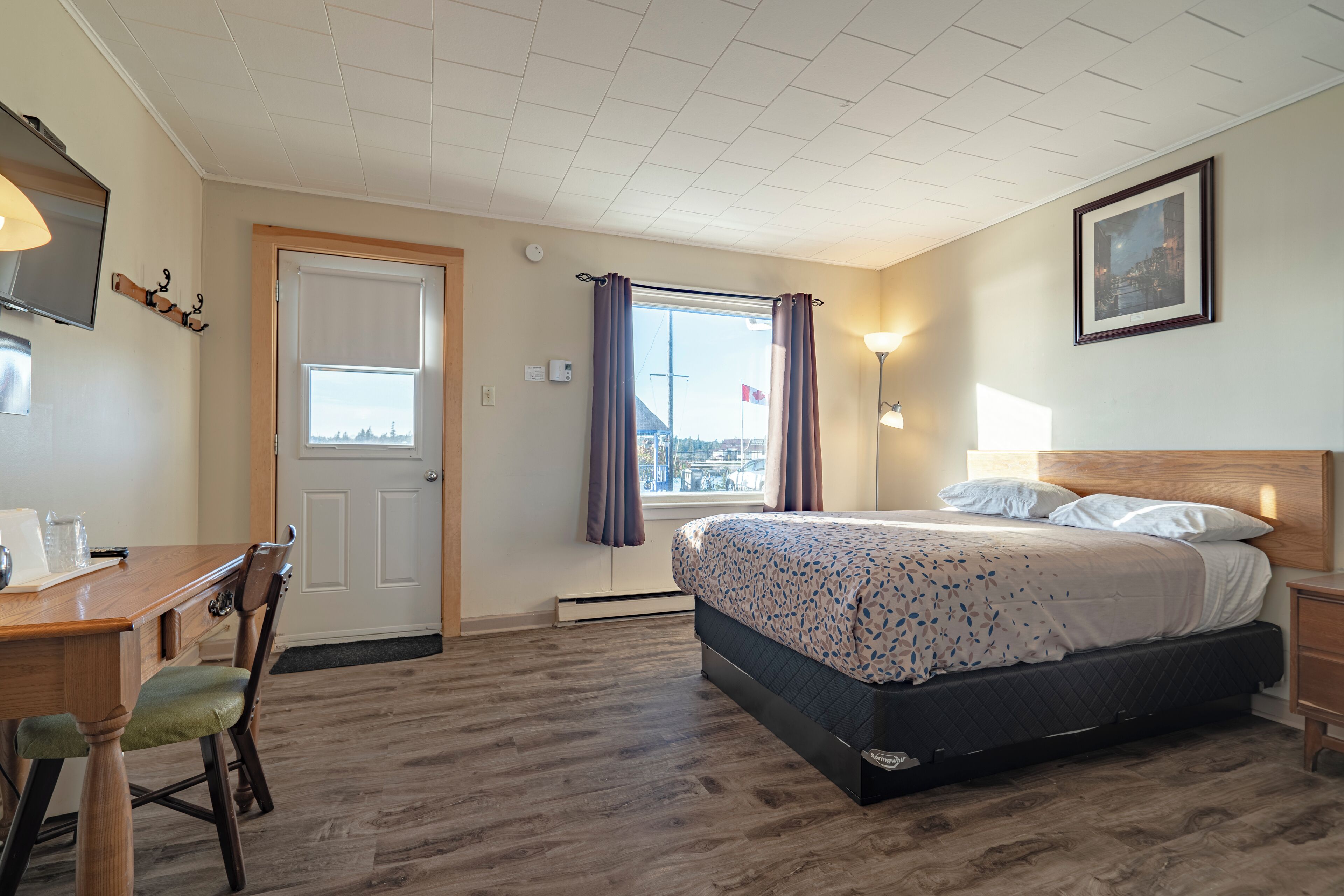 A hotel room with a bed, a desk, a chair, a window with curtains, a framed picture on the wall, and a ceiling with a grid pattern.