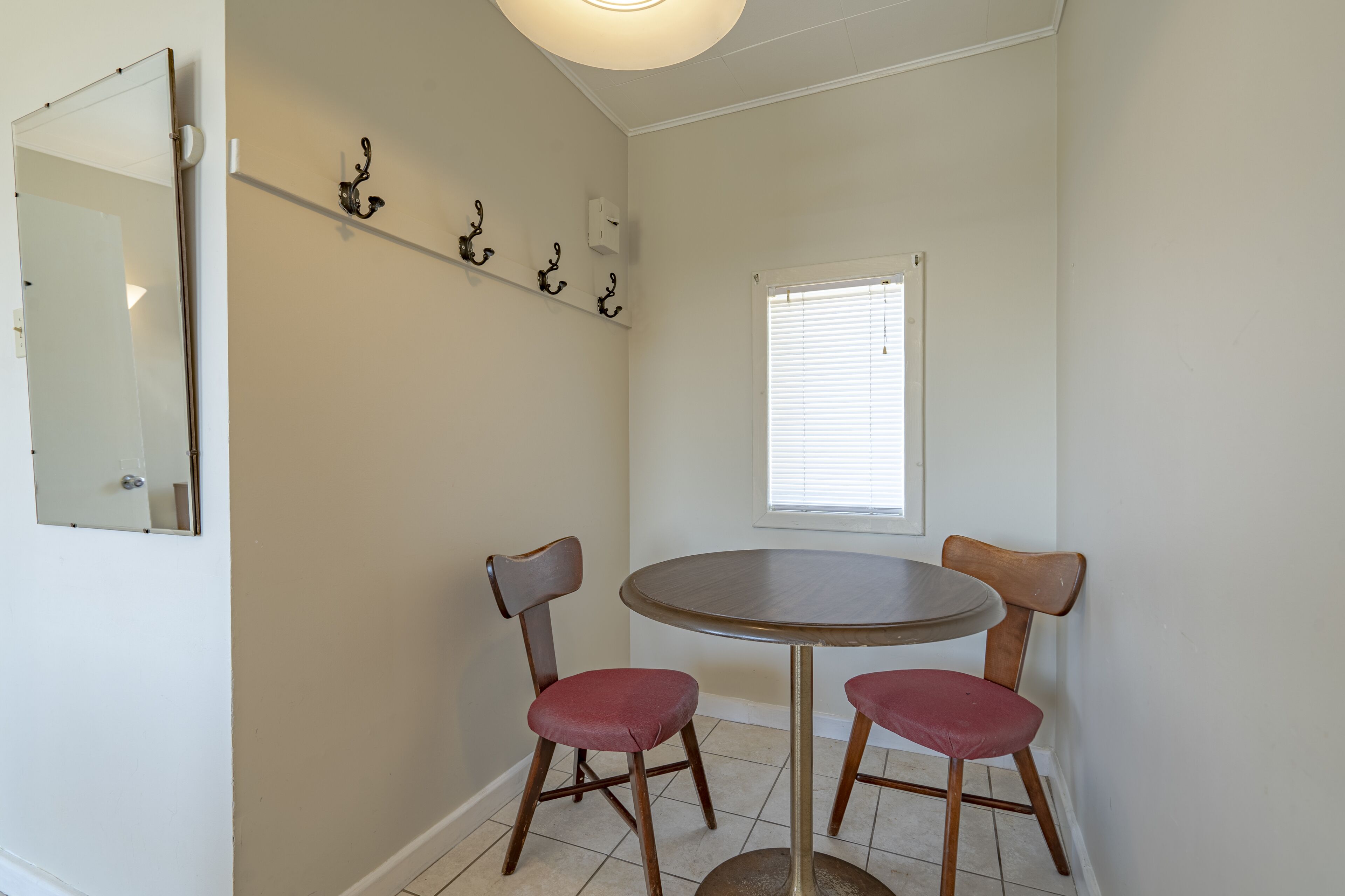 A dining area with a round table and two chairs, a wall-mounted shelf with hooks, a mirror, and a window with blinds.