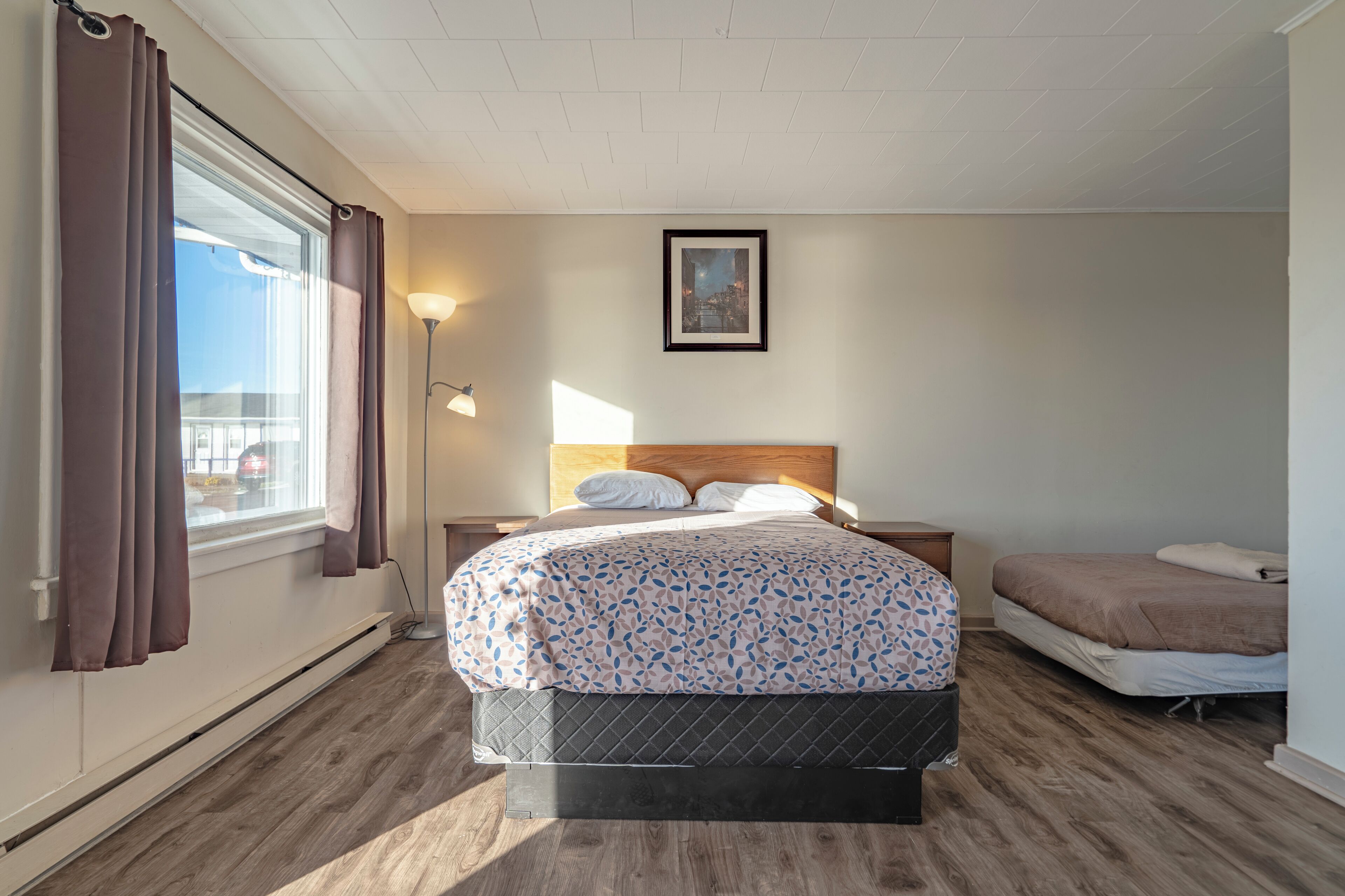 Un dormitorio con cama, mesita de noche, lámpara, ventana con cortinas y un cuadro en la pared.