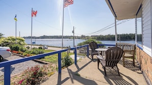 A covered patio with outdoor seating, overlooking a lake with flags, a parking area, and a view of a distant bridge. - NovaStar Motel On The Lake (Dayton)