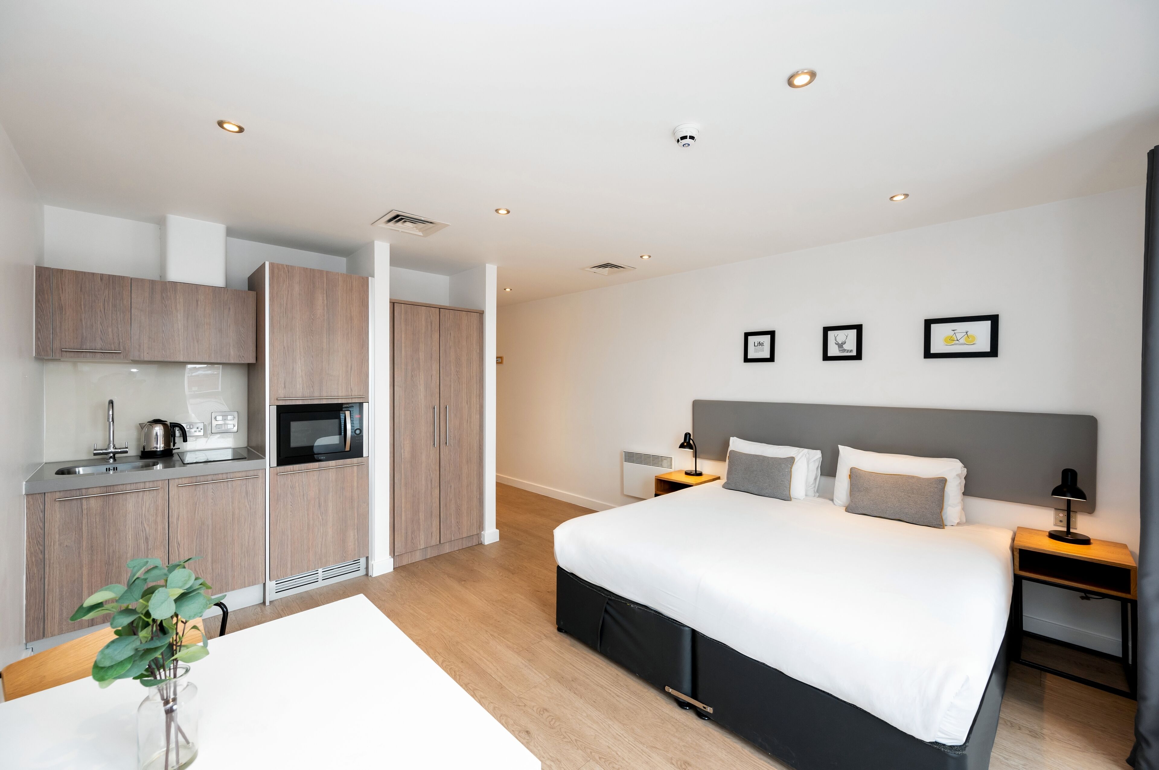 Una habitación de hotel moderna con una kitchenette independiente, una cama grande con cabecera gris, mesitas de noche con lámparas y una cámara de seguridad montada en la pared.