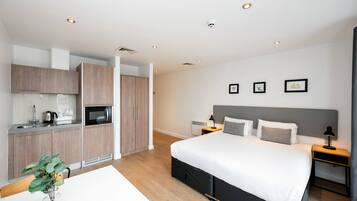 A modern hotel room with a separate kitchenette, a large bed with a grey headboard, bedside tables with lamps, and a wall-mounted security camera.