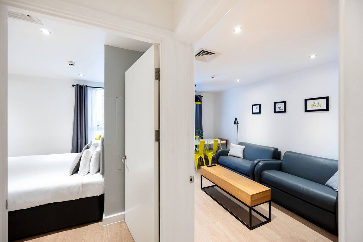 A modern hotel room with a bed, a sofa, a wooden bench, and a dining area with a yellow chair.