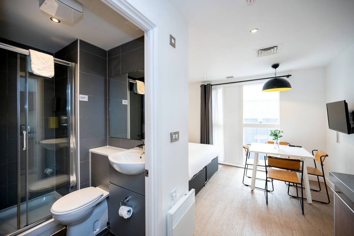 A modern bathroom with a glass shower enclosure, a white toilet, and a sink. There is a dining area with a table and chairs, and a large window providing natural light.