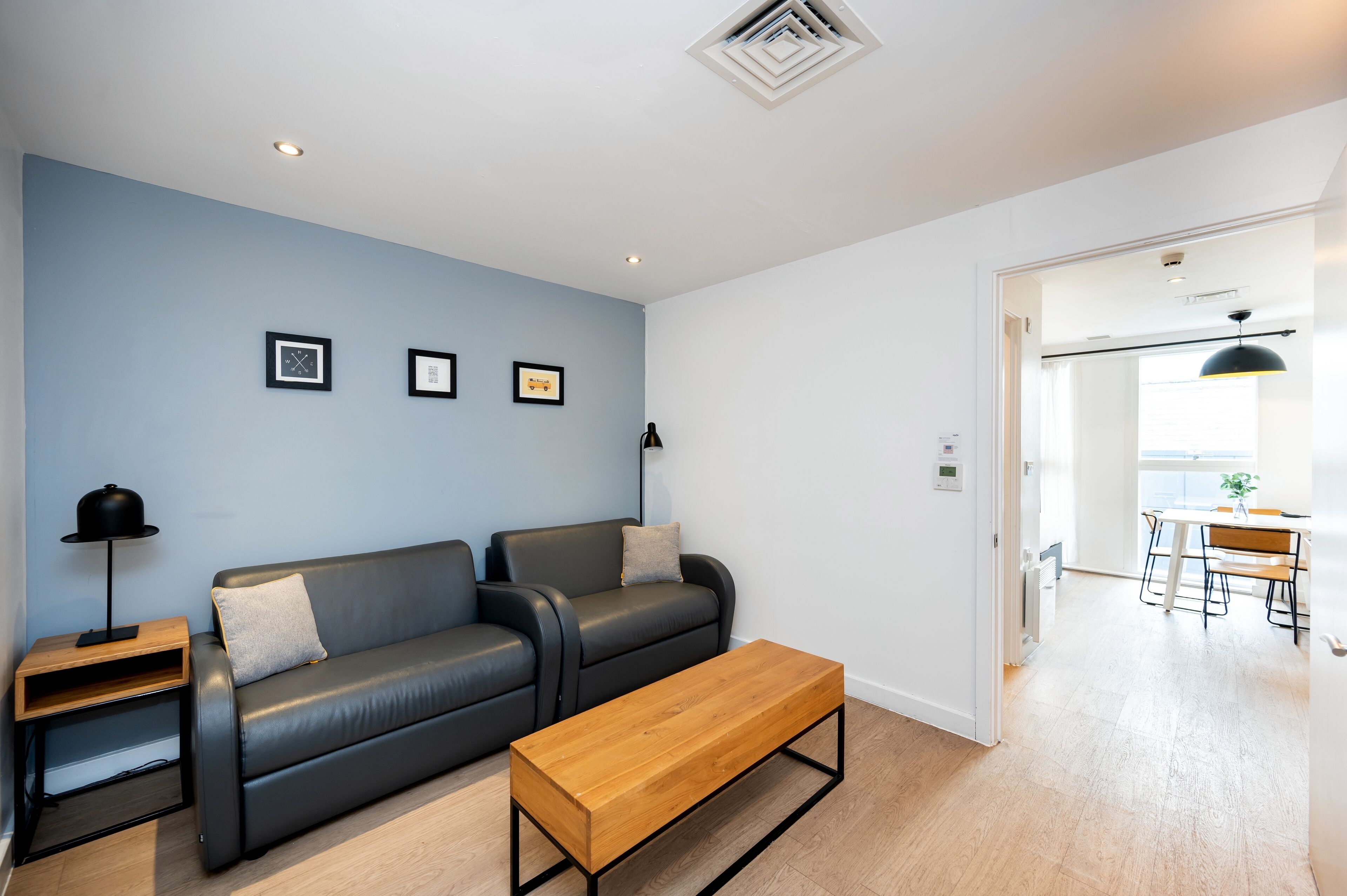 Una sala de estar moderna con un sofá de cuero negro, una mesa de centro de madera y una mesita de noche con una lámpara. Se observan cuadros en la pared y un área de comedor visible a través de una puerta abierta.