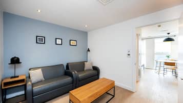 A modern living room with a black leather sofa, a wooden coffee table, and a side table with a lamp. There are framed pictures on the wall and a dining area visible through an open door.