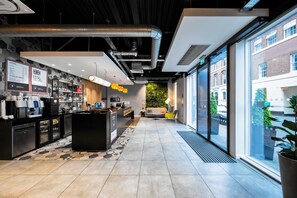Interior moderno de una cafetería con mostrador de recepción, zona de estar y grandes ventanales con vistas a la calle.