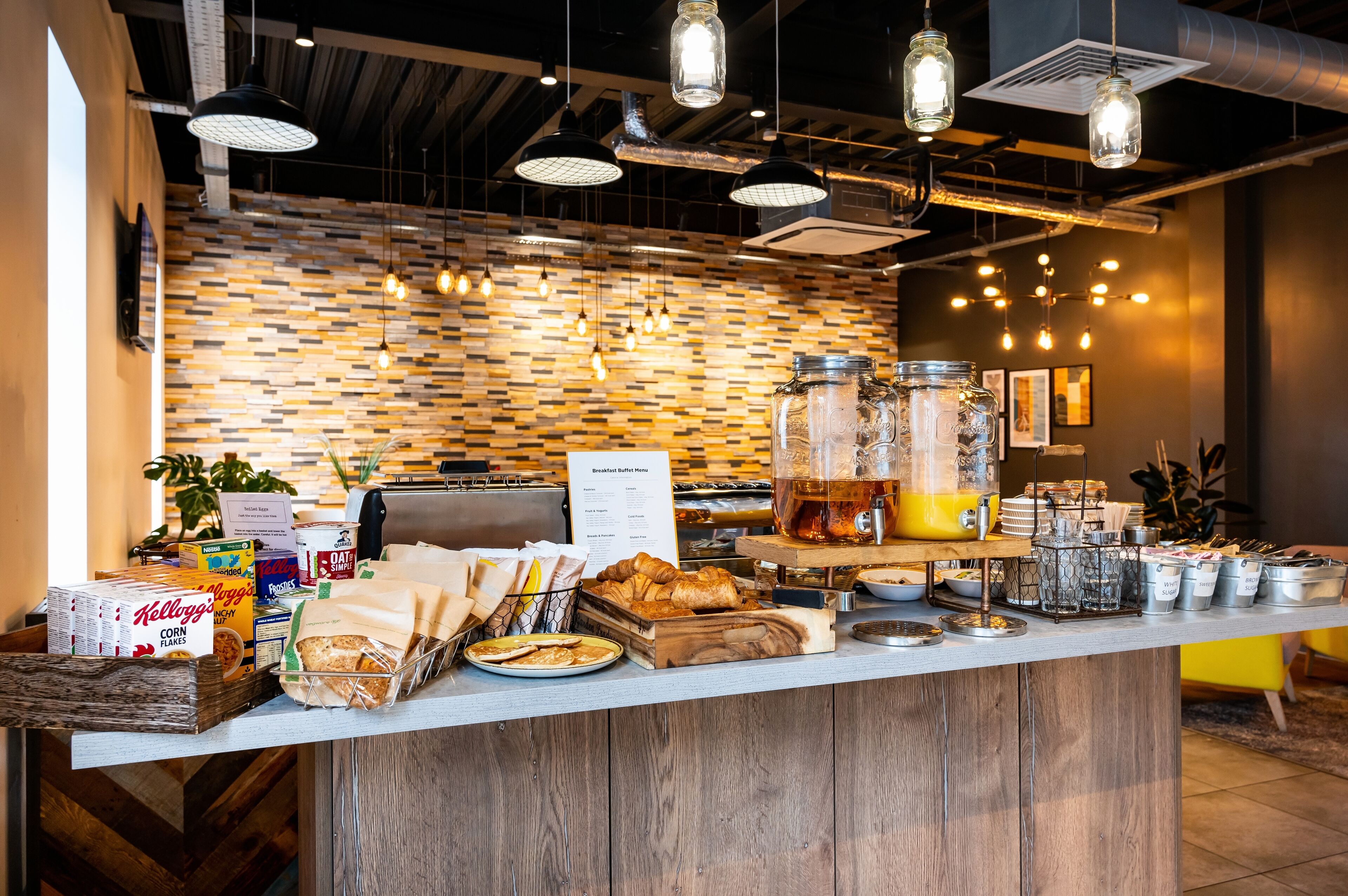 A breakfast buffet counter with various food items, a menu sign, and hanging light fixtures.