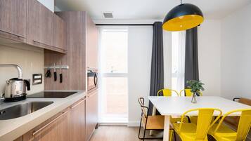 Cuina moderna amb armaris de fusta, taulell blanc i un brollador. Hi ha una zona de menjador amb taula blanca i cadires grogues.
