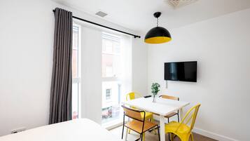 A modern hotel room with a large bed, a dining table with yellow chairs, a flat-screen TV mounted on the wall, and a window with curtains.