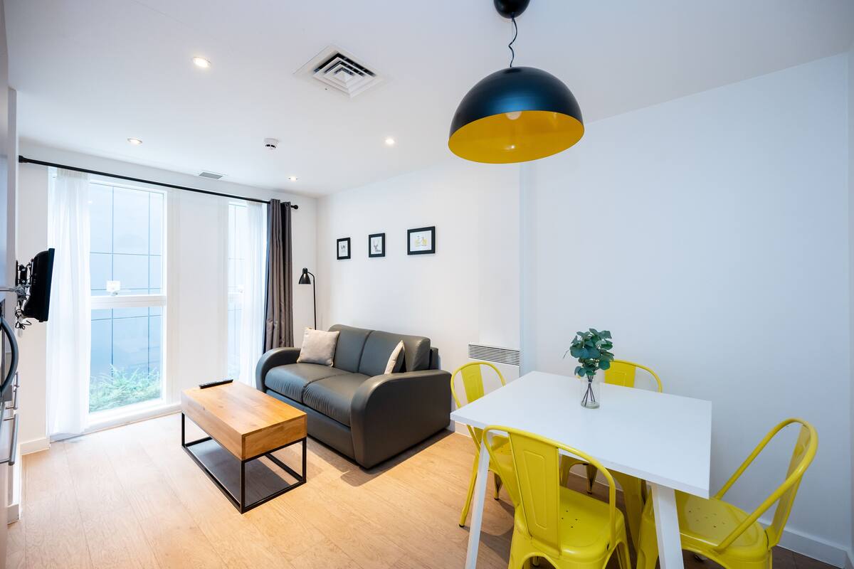 A modern living room with a sofa, a dining table with yellow chairs, and a large window with curtains.