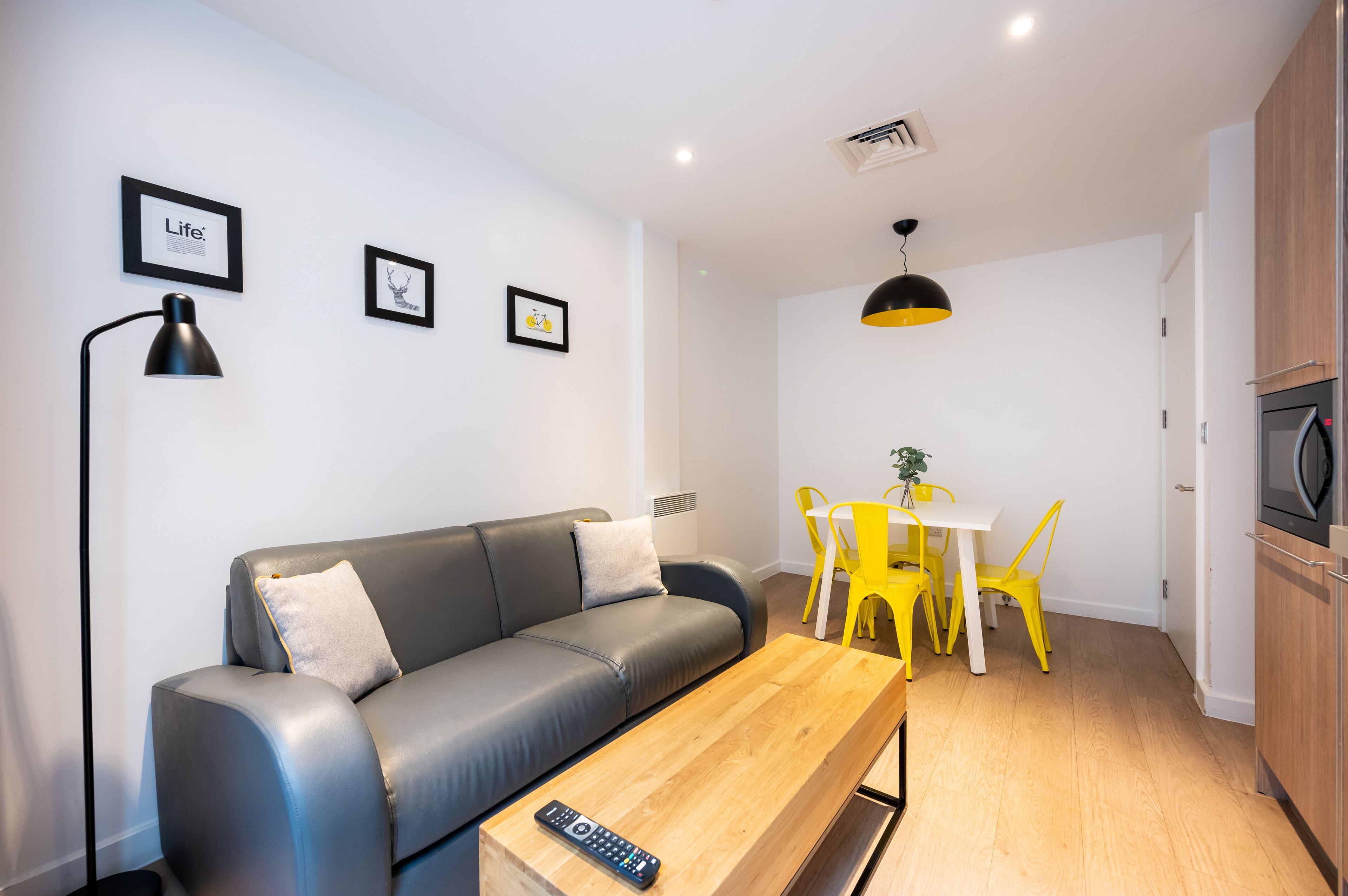 A modern living room with a grey sofa, a wooden coffee table, a remote control, a dining table with yellow chairs, and a kitchen area with wooden cabinets and a microwave.