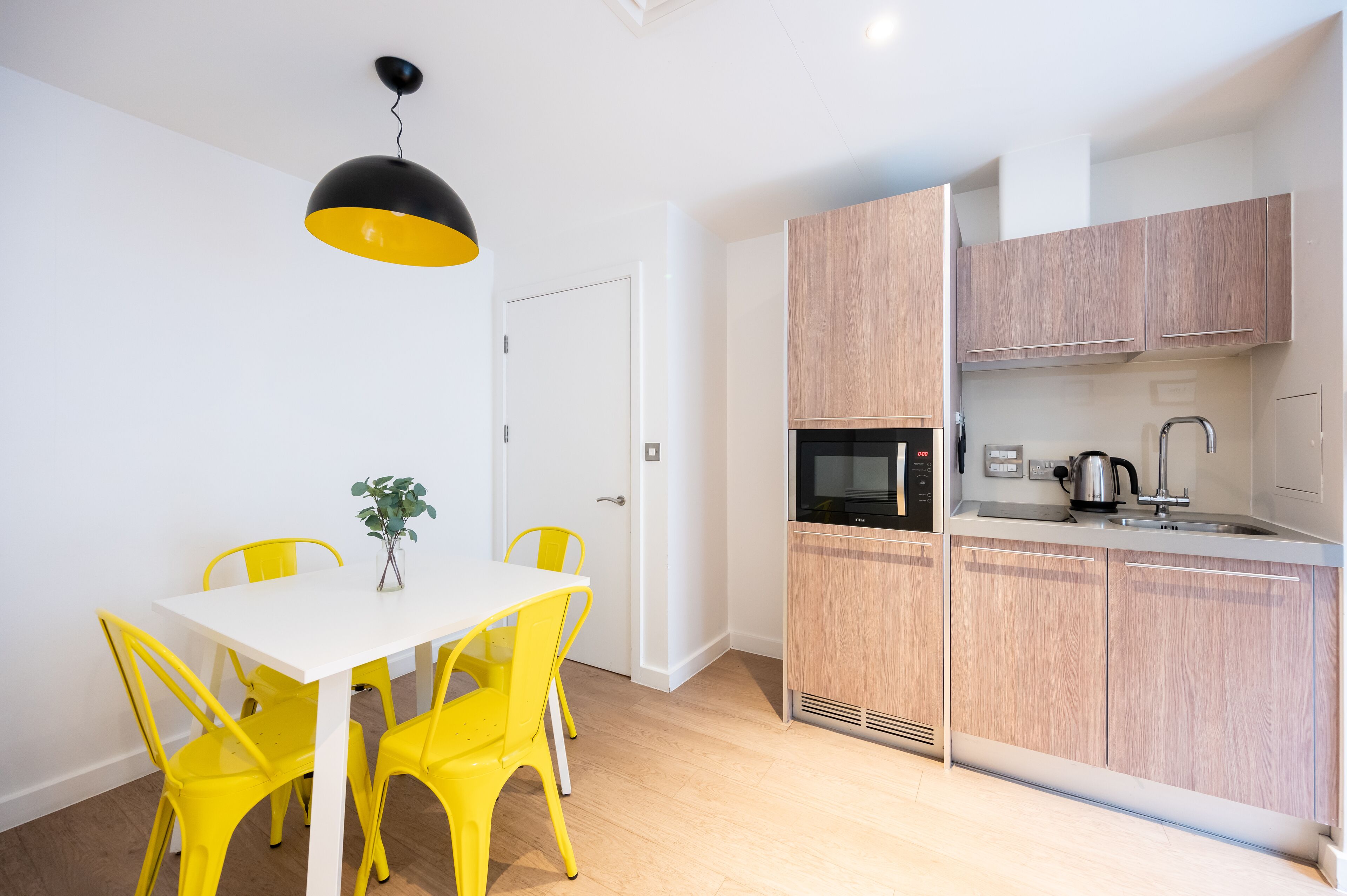 A modern kitchen with wooden cabinets, a microwave, and a kettle. There is a white dining table with yellow chairs and a black pendant light.