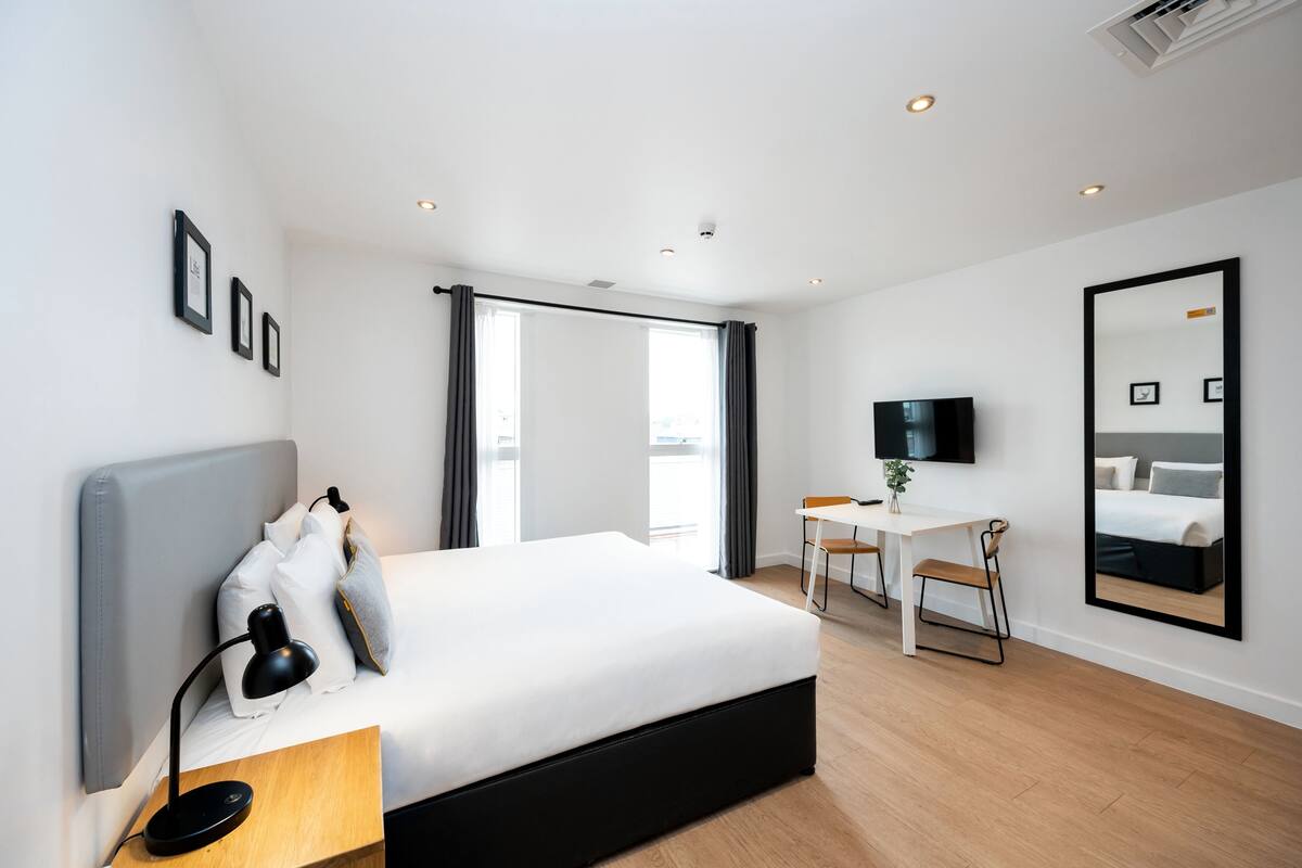 A modern hotel room with a large bed, a desk, a chair, a TV, and a large mirror on the wall.