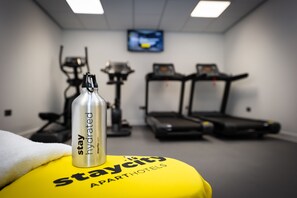 Un gimnasio con cintas de correr y equipo de ejercicio, una botella de agua sobre una toalla amarilla y un televisor en la pared.