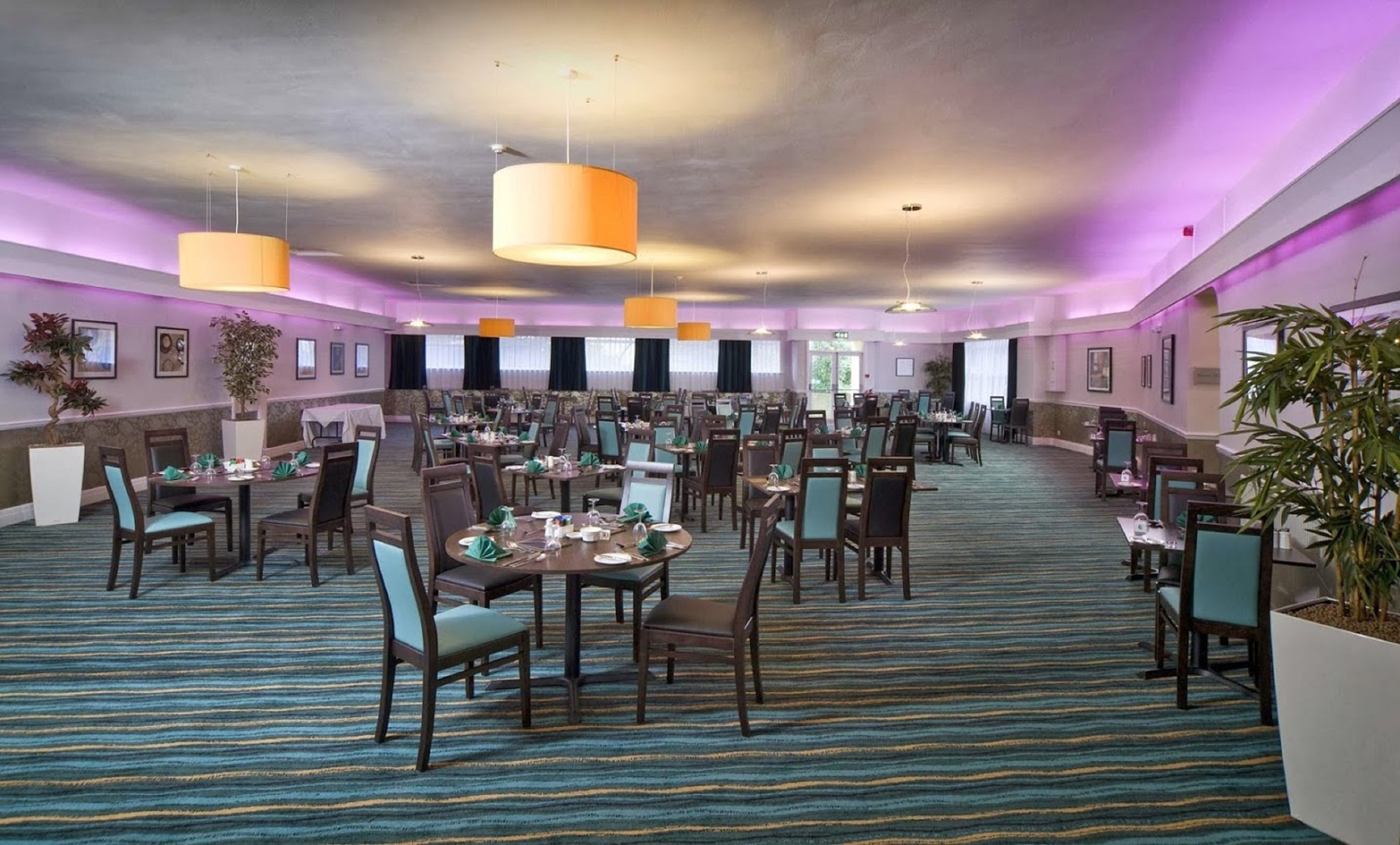 A large dining hall with multiple tables and chairs, ceiling lights, and potted plants.