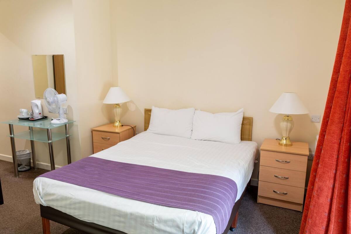 A hotel room with a bed, two bedside tables with lamps, a small desk with a fan, a mirror, and a trash bin.