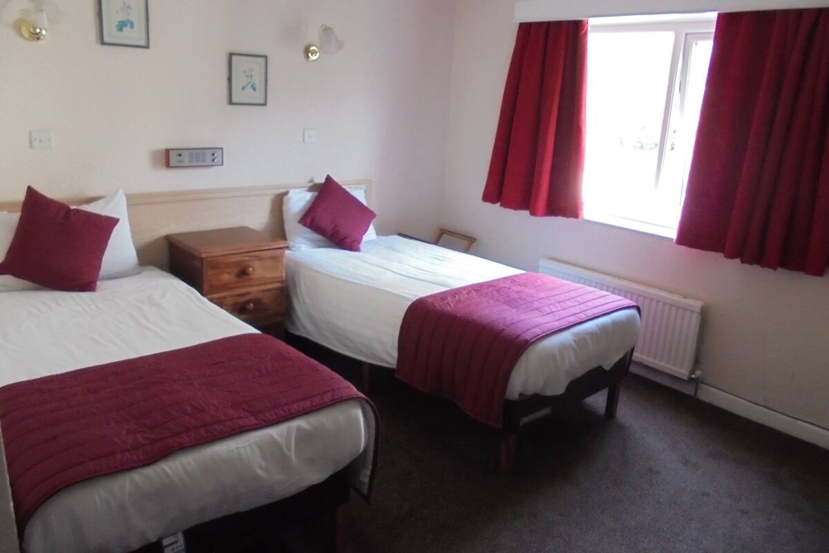 A room with two beds, each with a red blanket, a wooden nightstand, a white radiator, and a window with red curtains.