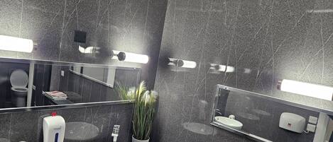 Une salle de bain moderne avec un grand miroir, un lavabo blanc, un sèche-mains et une plante en pot.