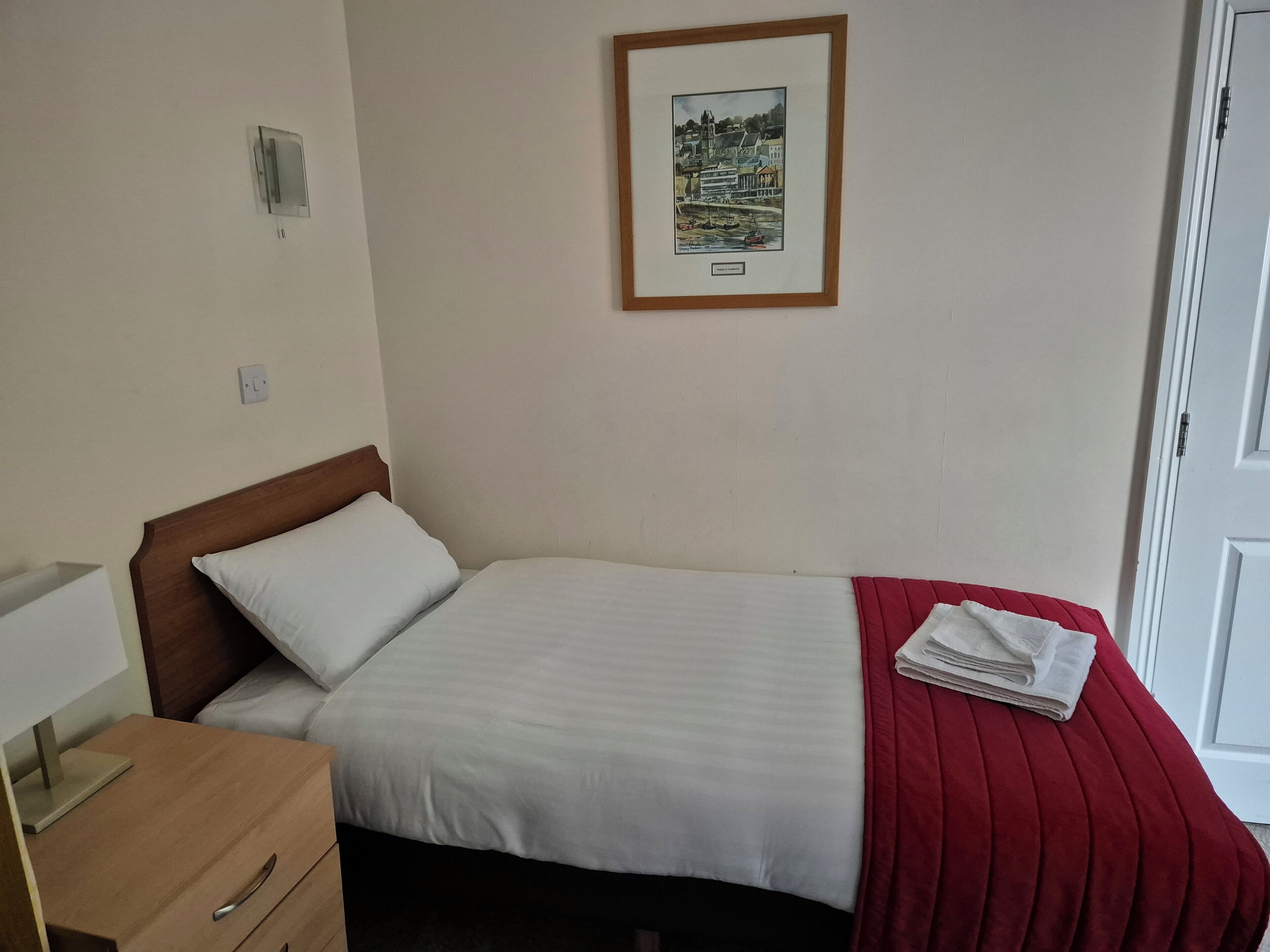 Un lit bien fait, recouvert d’une couverture blanche et rouge, avec une tête de lit en bois, une table de chevet sur laquelle se trouve une lampe, et un tableau encadré accroché au mur.