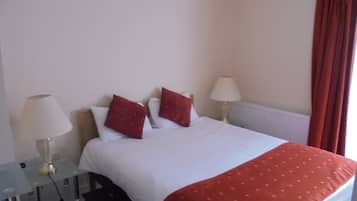 A hotel room with a bed, two bedside lamps, a red throw blanket, and a window with red curtains.