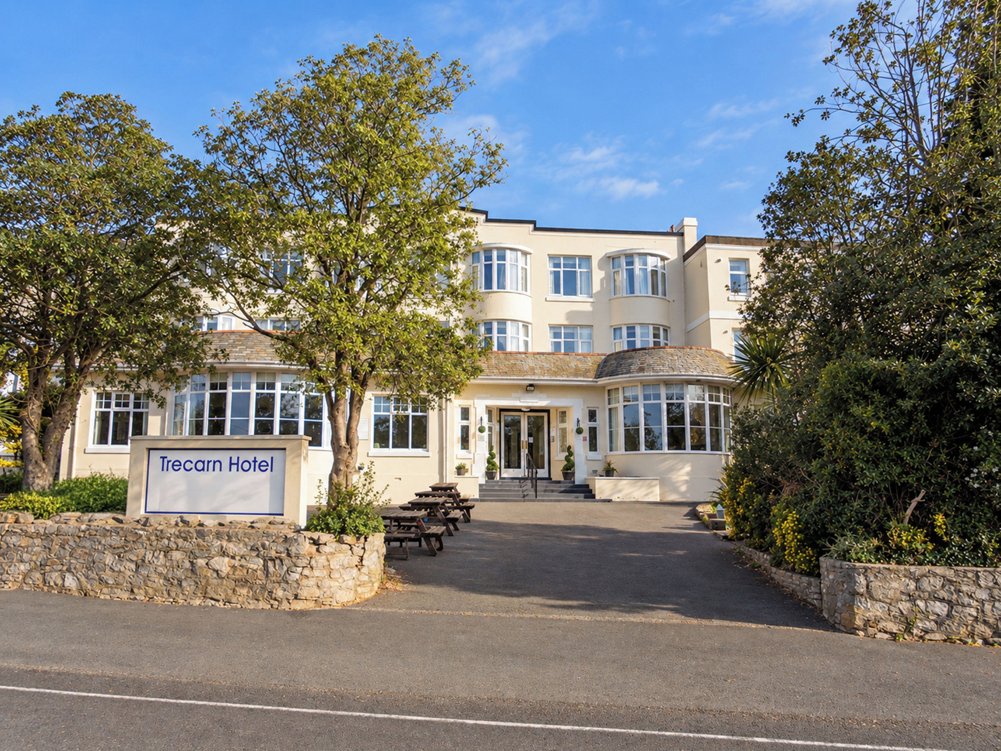 A hotel with a sign reading 'Trecarn Hotel,' a paved entrance, and a stone wall with greenery.