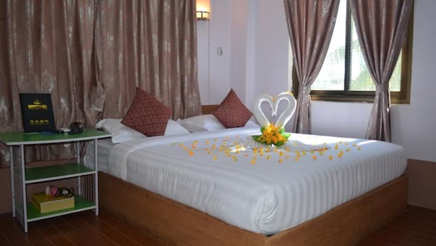Una cama bien hecha con sábanas blancas, un arreglo floral en forma de corazón y pétalos de rosa esparcidos. Hay una mesita con un objeto negro encima, y una estantería verde debajo.