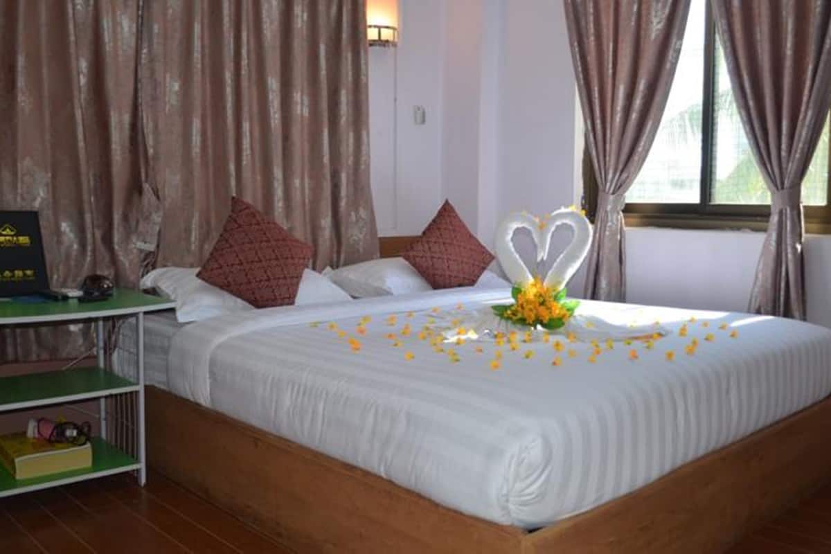 Una cama bien hecha con sábanas blancas, un arreglo floral en forma de corazón y pétalos de rosa esparcidos. Hay una mesita con un objeto negro encima, y una estantería verde debajo.