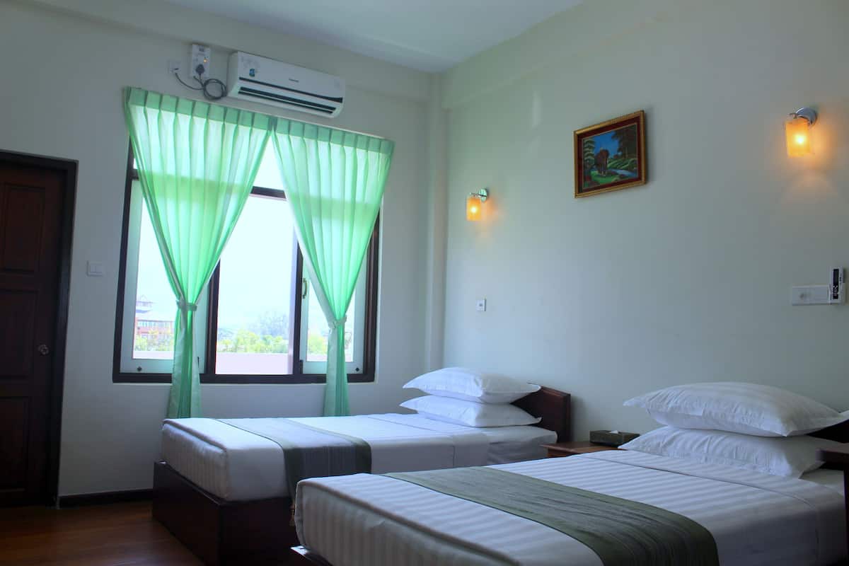 A hotel room with two beds, a window with green curtains, an air conditioning unit, a wall-mounted light, and a framed picture.