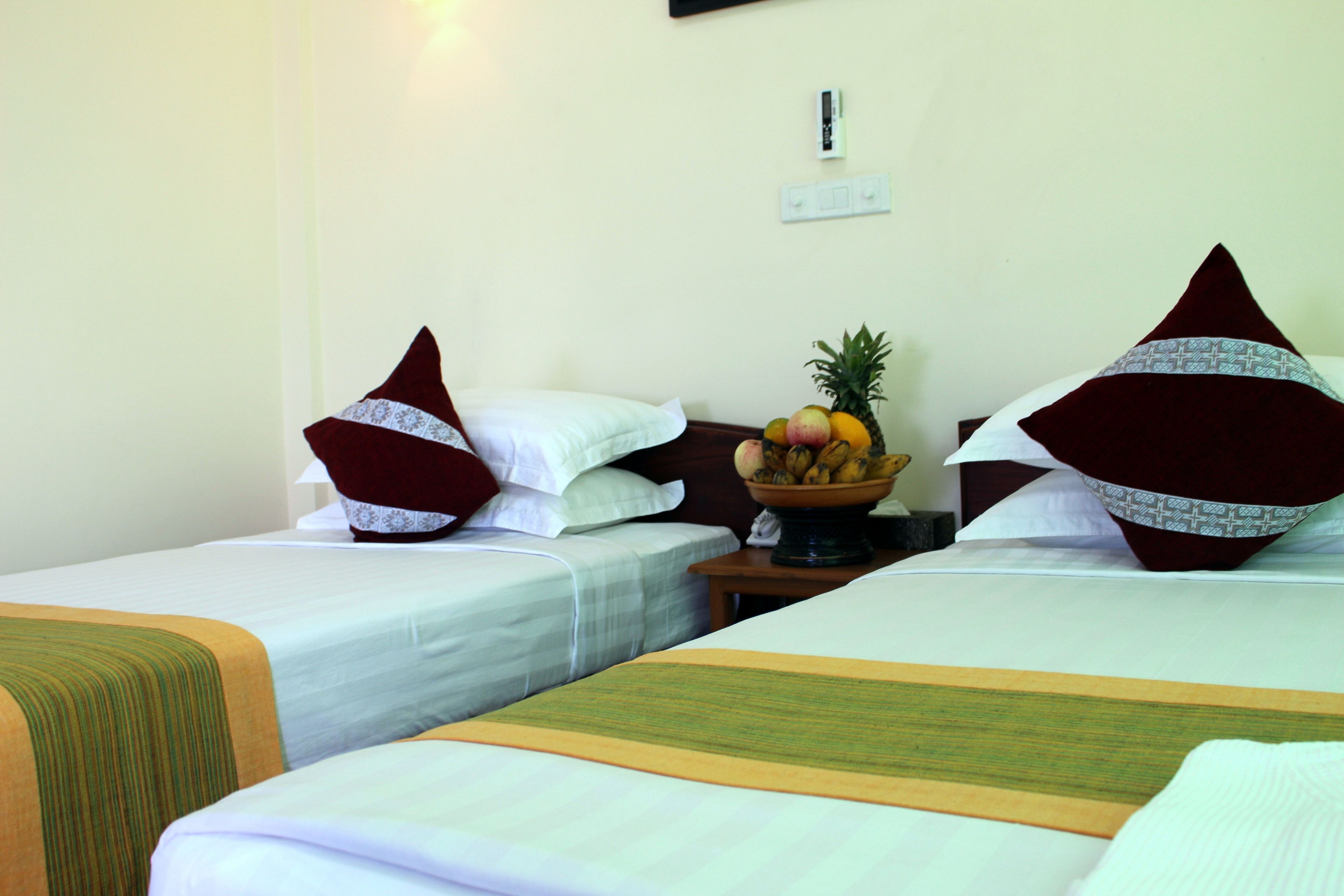 A hotel room with two single beds, each with a pillow and a bedside table. There is a fruit bowl on the nightstand.