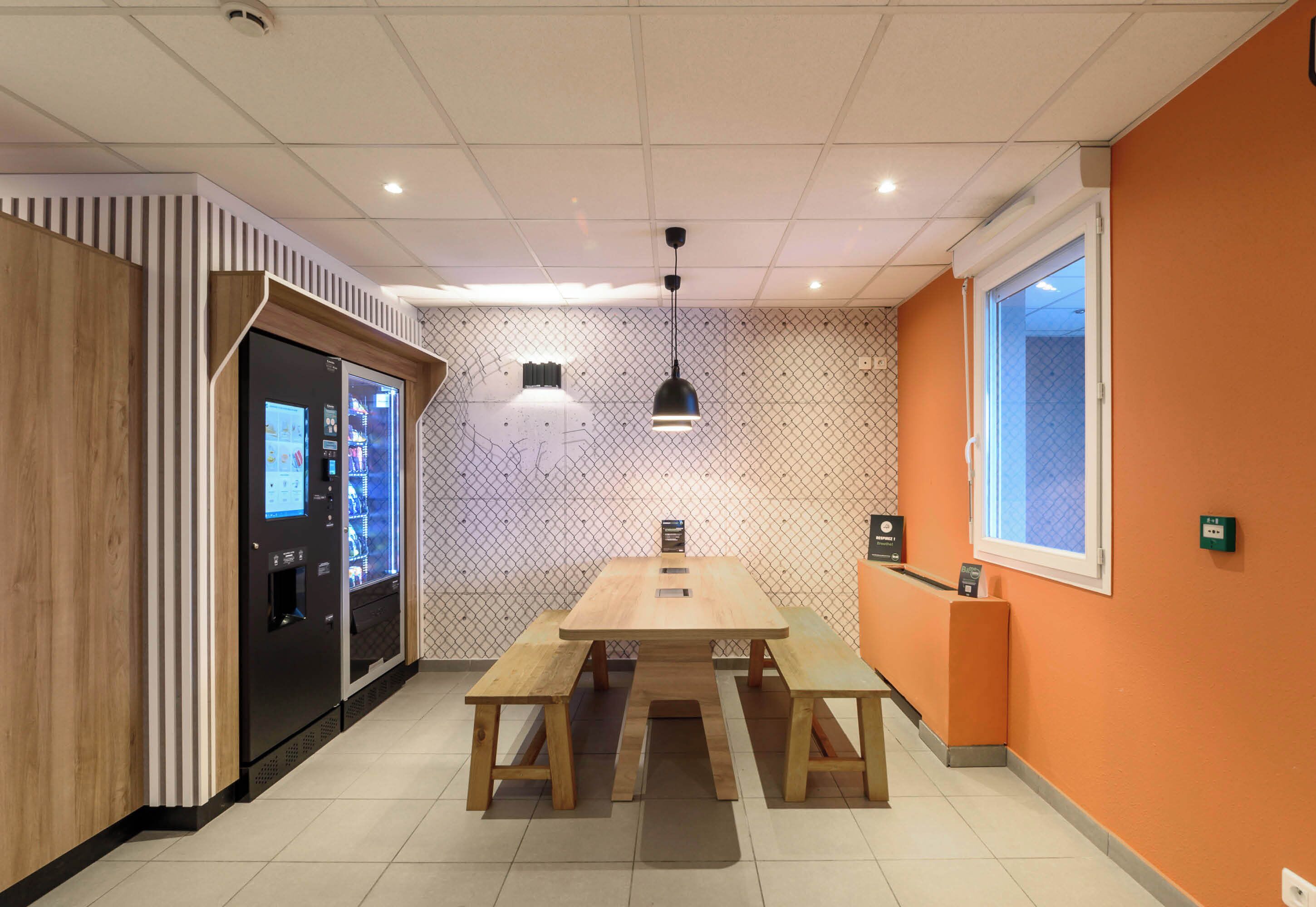 Un espacio interior moderno con una mesa de madera y bancos, una máquina expendedora y un muro con estampado.