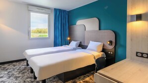 Habitación de hotel con dos camas, un ventanal con cortinas, un escritorio y una lámpara fijada a la pared.