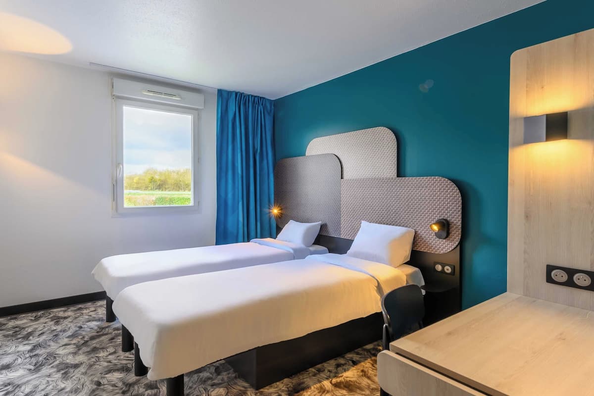 Habitación de hotel con dos camas, un ventanal con cortinas, un escritorio y una lámpara fijada a la pared.