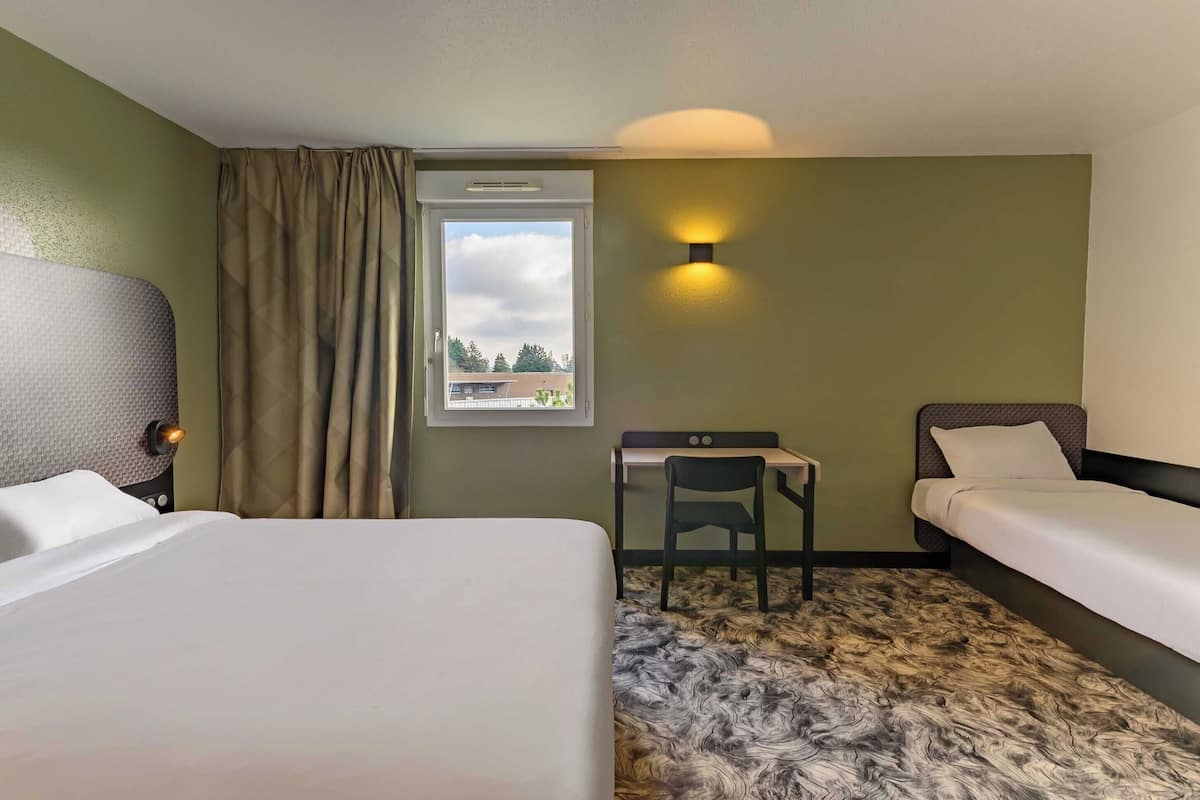 Habitación de hotel con dos camas, un escritorio, una silla, un ventanal con cortinas y una lámpara en la pared.