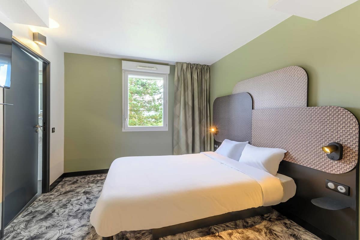 Habitación de hotel con cama, ventana con cortinas, luz empotrada en la pared y puerta.