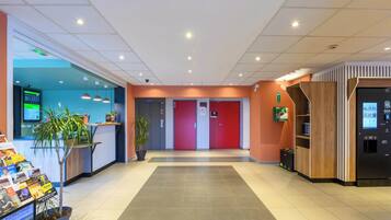 A modern hotel lobby with a reception desk, a digital information board, a vending machine, and a potted plant.
