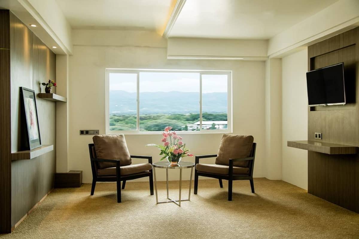 Una habitación con dos sillones, una pequeña mesa con un jarrón de flores, un amplio ventanal con vistas a montañas y un televisor de pantalla plana montado en la pared.