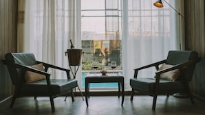 A room with two armchairs, a small table with a bottle and glasses, and a view through a window with sheer curtains. - Hotel Hazel (Mandalay)