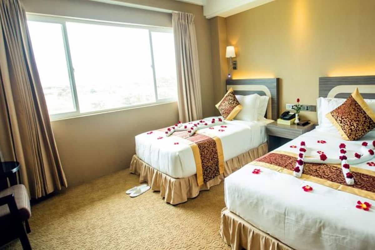 Habitación de hotel con dos camas, una mesita de noche, una lámpara y vistas al exterior.