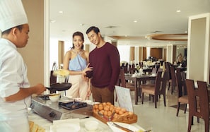 A dining area with tables and chairs, a buffet with eggs, and a person serving food. - Hotel Hazel (Mandalay)