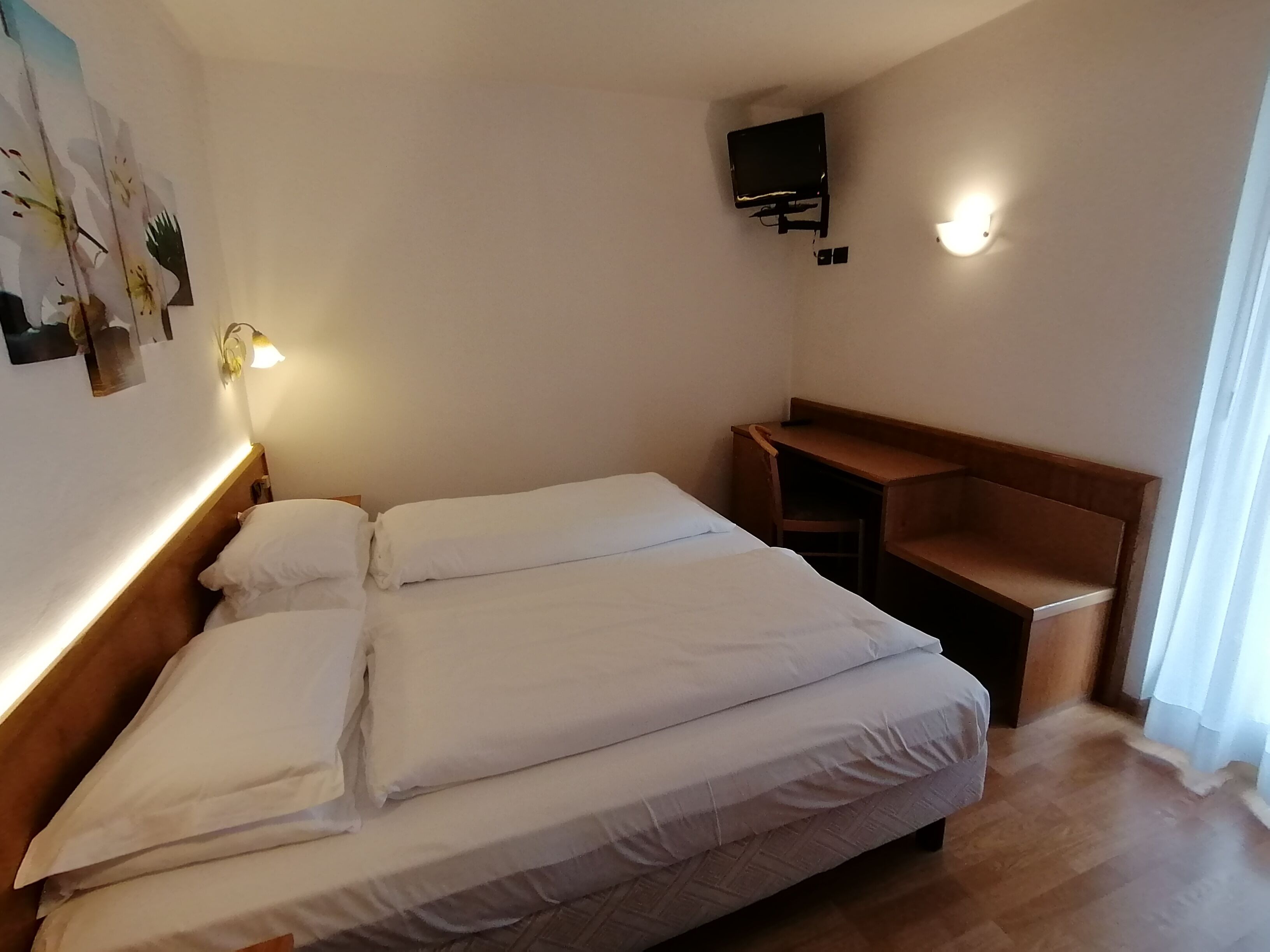 A hotel room with a bed, a desk, a wall-mounted TV, and a light fixture.