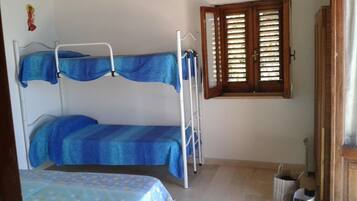 A small, simple room with a bunk bed, a window with wooden shutters, a cross on the wall, and a small cabinet.