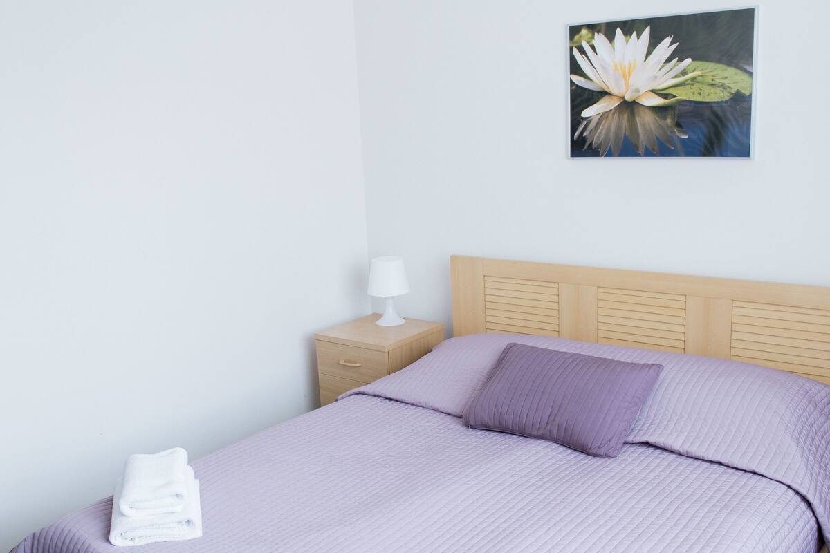 Una cama bien tendida con una colcha morada y dos almohadas, una lámpara de noche blanca, una mesita de noche de madera pequeña y un cuadro con una flor en la pared.