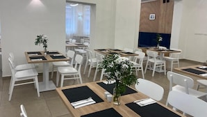 Un comedor moderno con sillas y mesas blancas, algunas con manteles individuales negros. Hay plantas en macetas sobre las mesas, y las paredes son lisas con un panel decorativo de madera.
