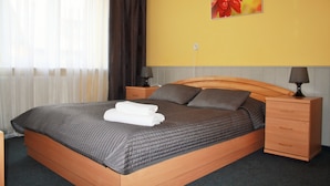 Una cama bien hecha con cabecera y piecera de color marrón oscuro, una mesita de noche de madera con una lámpara, y un cuadro de flores rojas colgado en la pared.