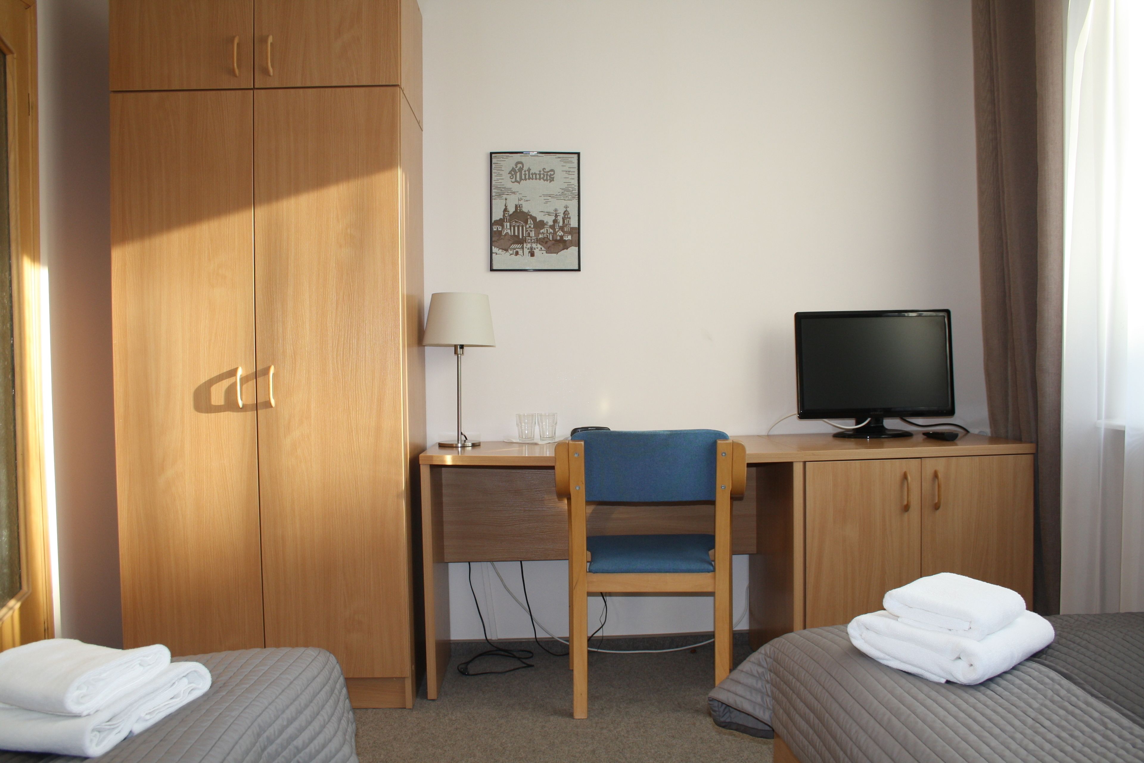 Quarto de hotel com escrivaninha de madeira, uma cadeira, cama com duas almofadas, televisão e quadro pendurado na parede.