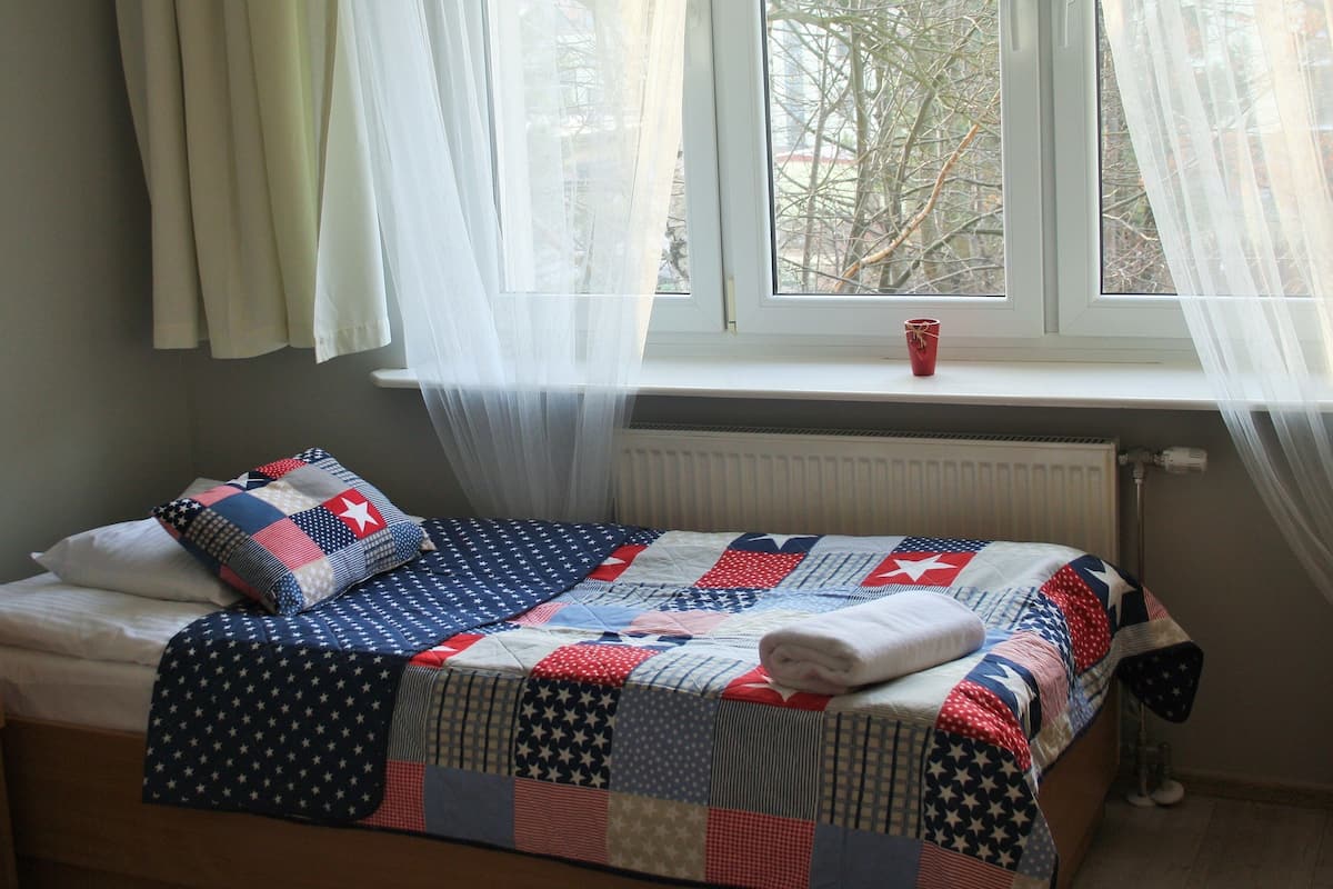 Una cama individual con colcha, una ventana con cortinas y un radiador.