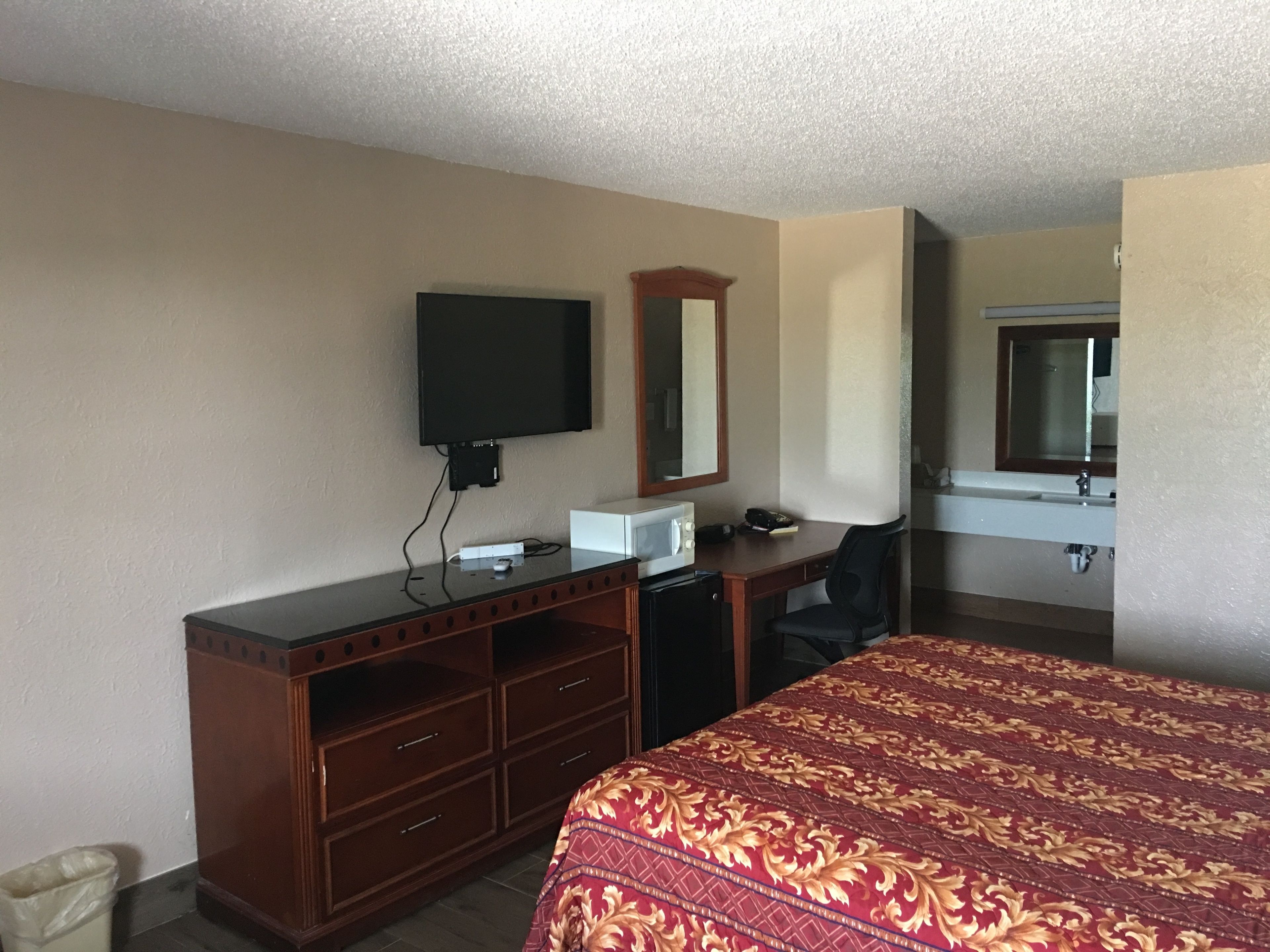 A hotel room with a bed, a desk with a computer, a TV, a mirror, and a bathroom with a sink and mirror.