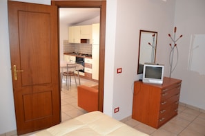 Une chambre avec une commode en bois, un téléviseur posé dessus, et une porte donnant sur un coin cuisine.
