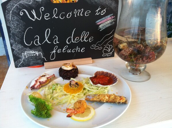 Prato de mariscos e vegetais, com uma placa indicando 'Welcome to Cala delle Feluche' ao fundo.